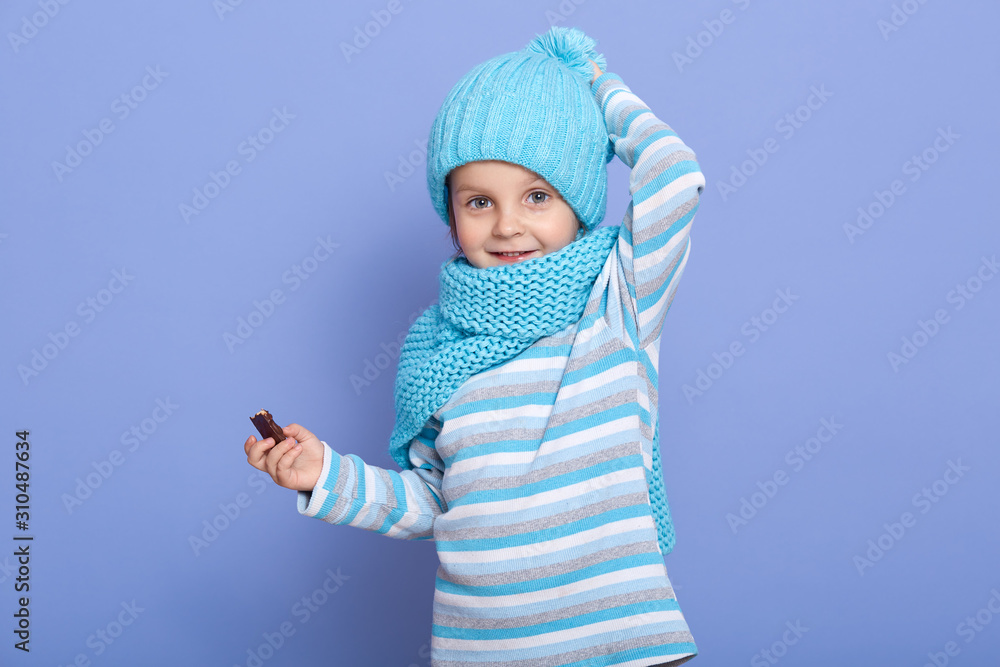 Charming astionish little girl looking at camera with surprised facial expression isolated over blue studio background, child holding candy in hand and keeps hand on head, wears cap, scarf and shirt.