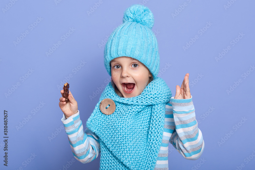 Close up portrait of little girl wearing winter warm turquoise cap and scarf posing over purple background, female child with opened mouth, looks astonished, being shocked, has pleasant look. Close up portrait of little girl wearing winter warm turquoise cap and scarf posing over purple background, female child with opened mouth, looks astonished, being shocked, has pleasant look.