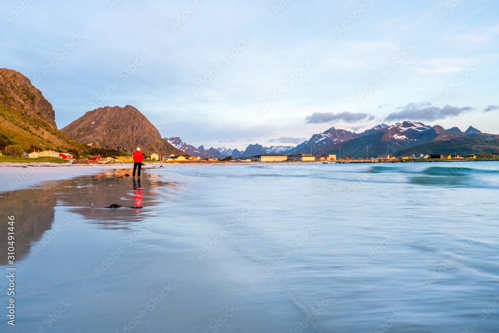 Fototapeta premium Man in red jacket stands on Ramberg beach in Lofoten island in Norway during sunset. Houses and mountains in the background. Travelocity and traveling concept.
