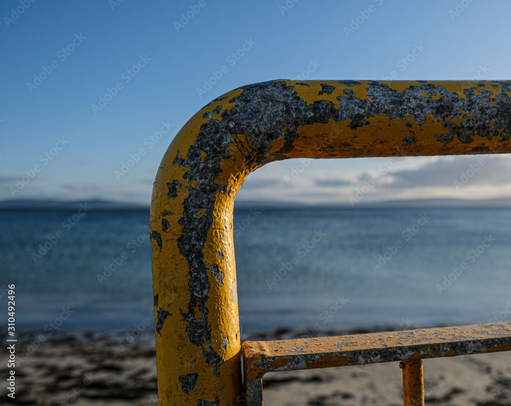 Fototapeta premium Rusty yellow railings along the beach on Salthill Promenade, near Gawlay in Ireland, with the waters of the bay behind.
