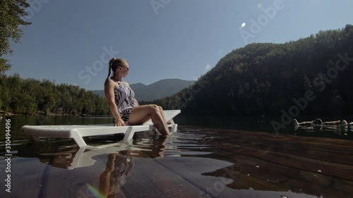 Woman lie on a sunbed in sunglasses and a boho silk shawl. Girl rest on a flood wood underwater pier. The pavement is covered with water in the lake. In the background are mountain and a forest. photo