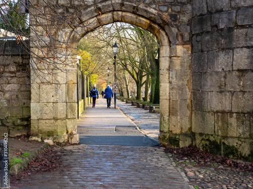 View through a stone arch to a couple strolling on the tree lined riverside Dame Judi Dench Walk in York, England photo
