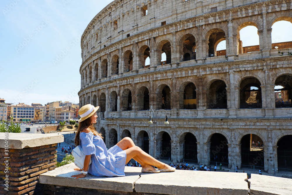 Travel woman in romantic dress and hat sitting and looking on Coliseum, Rome, Italy. Beautiful tourist girl with backpack near Colosseum. Young woman enjoy summer Italian vacation in Europe.