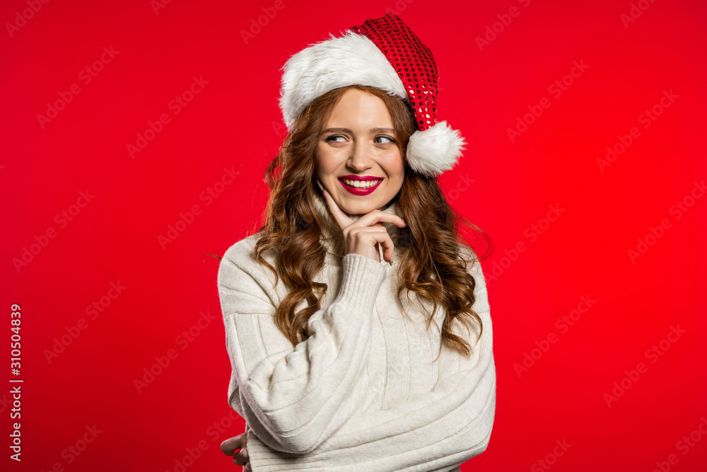 Portrait of attractive woman with sly smile, red lips in Santa hat and warm sweater on studio background. Girl in good winter christmas mood. Portrait of attractive woman with sly smile, red lips in Santa hat and warm sweater on studio background. Girl in good winter christmas mood.