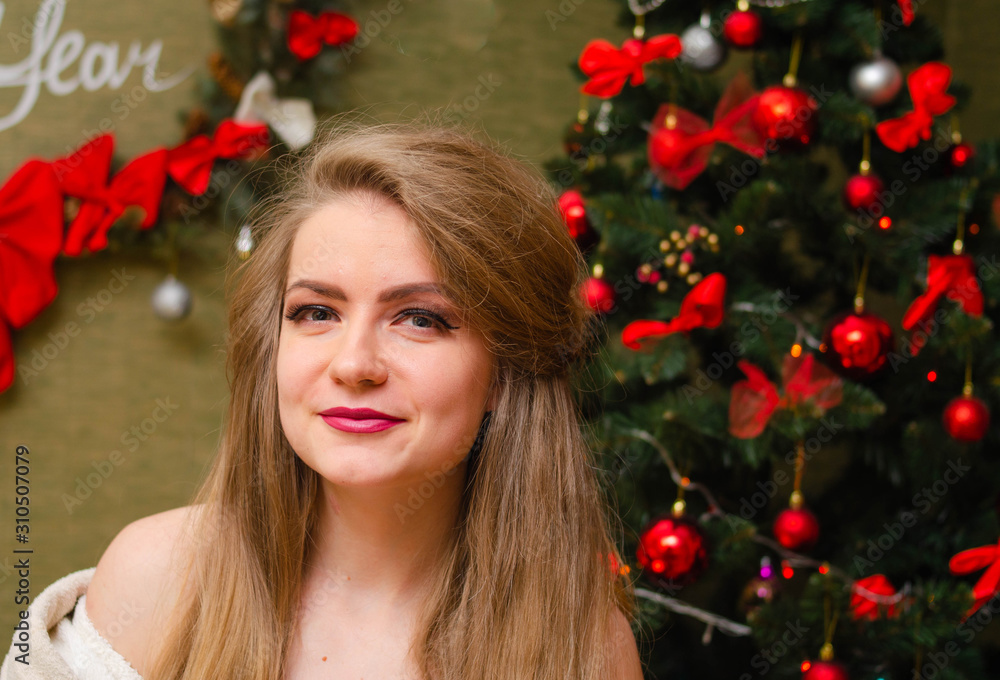 Portrait of a girl with bright red lips, blond long hair against the background of a New Year tree. Young girl in a white warm coat, shoulders are visible. Holidays. Merry Christmas. Portrait of a girl with bright red lips, blond long hair against the background of a New Year tree. Young girl in a white warm coat, shoulders are visible. Holidays. Merry Christmas.