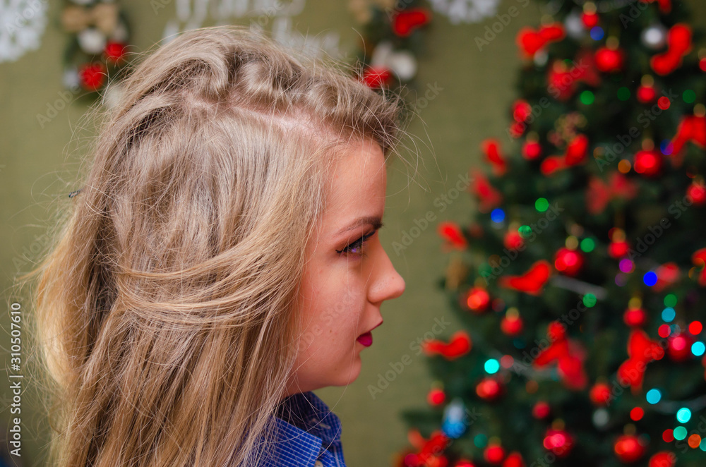 Portrait of a girl with bright red lips, blond long hair against the background of a New Year tree. Young girl in a blue men's shirt. Holidays. Merry Christmas. funny Face.