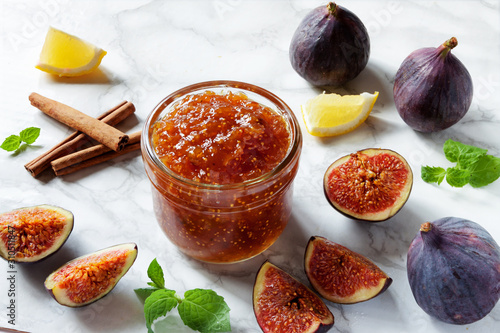 Fresh figs, with a dish of homemade fig jam or preserves with lemon cinnamon sticks and mint on white kitchen marble table background. Healthy breakfast dessert snack recipes and veggie tables concept