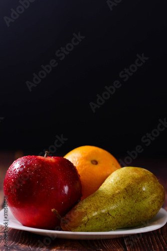 Fresh organic fruit rd apple  orange and pear in white plate on rustic wooden table. glossy fruit with black background. Vertical