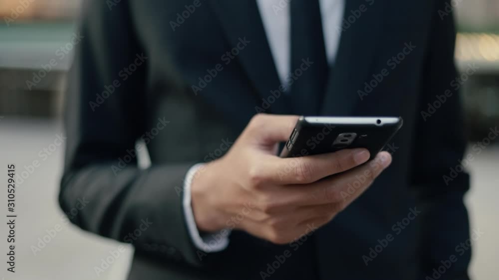 Handsome asian businessman in a black suit using his smartphone browsing internet, scrolling social media while standing outside on the street near a big office building urban having sunlight sunset. Handsome asian businessman in a black suit using his smartphone browsing internet, scrolling social media while standing outside on the street near a big office building urban having sunlight sunset.