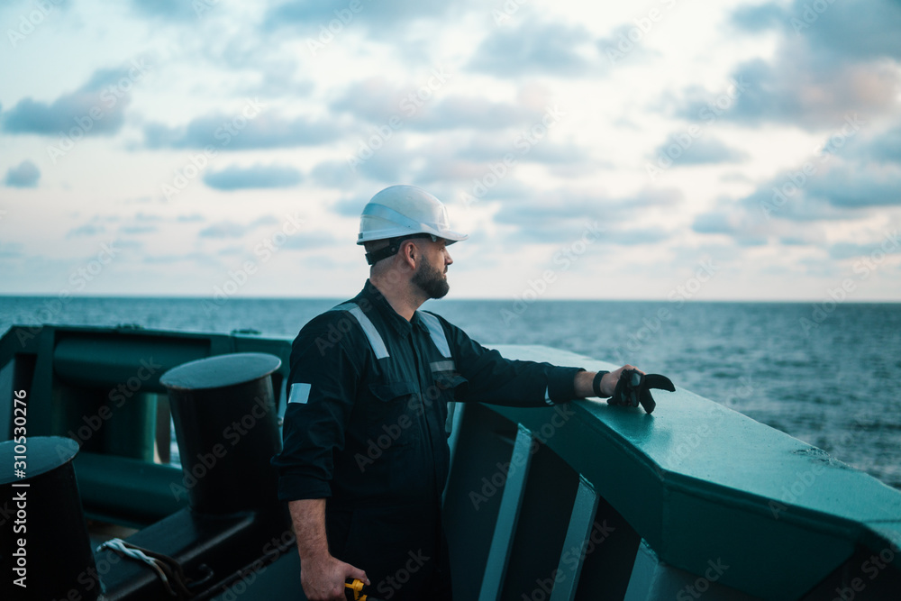 Obraz premium Marine Deck Officer or Chief mate on deck of offshore vessel or ship , wearing PPE personal protective equipment - helmet, coverall. He holds VHF walkie-talkie radio in hands.