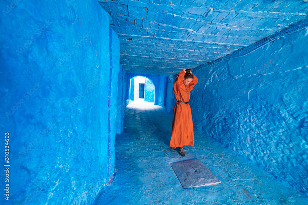 a woman in a bright orange long dress collects hair at the top of her head with her hands in the corridor of the blue city of Morocco a woman in a bright orange long dress collects hair at the top of her head with her hands in the corridor of the blue city of Morocco