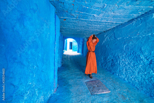 a woman in a bright orange long dress collects hair at the top of her head with her hands in the corridor of the blue city of Morocco © nelen.ru