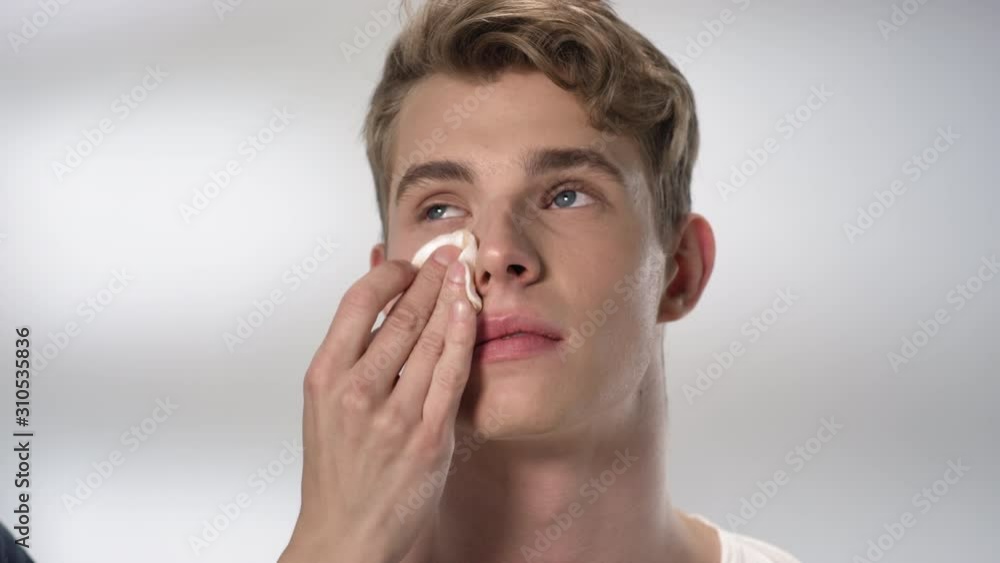 Lockdown of face of young Caucasian man and male visagiste hand holding cotton pad with cleansing tonic and wiping it Lockdown of face of young Caucasian man and male visagiste hand holding cotton pad with cleansing tonic and wiping it