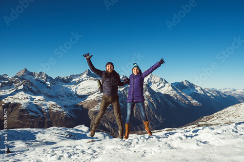 Happy travelers couple man and woman trekking in mountains, enjoy beautiful view. Explorers hiking on hills, travel in Swiss Alps, Switzerland. Hikers jumping with raised hands outdoors on nature.