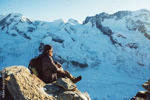 Traveler Man with backpack trekking in mountains, enjoy beautiful Matterhorn view. Explorer man hiking on snowy hills, travel in Alps, Switzerland. Hiker sitting on rock cliff outdoors on nature.