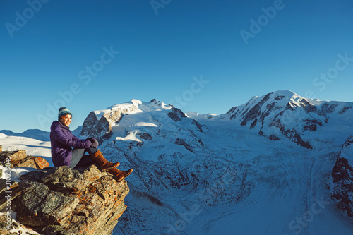 Happy traveler woman trekking in mountains, enjoy beautiful Matterhorn view. Explorer girl hiking on snowy hills, travel in Alps, Switzerland. Hiker sitting on rock cliff outdoors on nature.