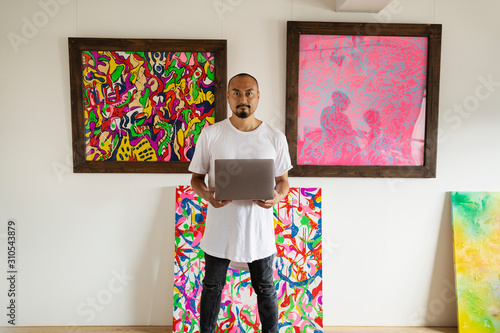 Japanese man standing in art gallery, holding laptop computer, looking at camera. photo