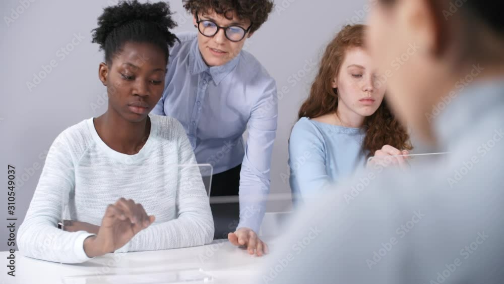Medium shot of Caucasian female teacher in glasses helping young Afro-American college student perform practical task on transparent touchscreen augmented reality device. Suitable for AR, VR effects.