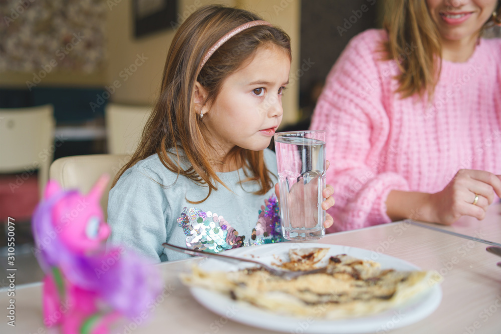 Portrait of small cute little girl caucasian sitting by the table at home or restaurant eating pancake dink water hold big glass Portrait of small cute little girl caucasian sitting by the table at home or restaurant eating pancake dink water hold big glass