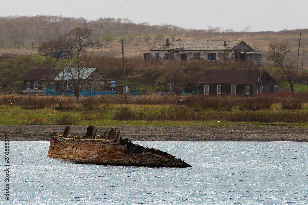 A wrecked flooded wooden ship sticks out of sea water against the backdrop of an island with wooden houses