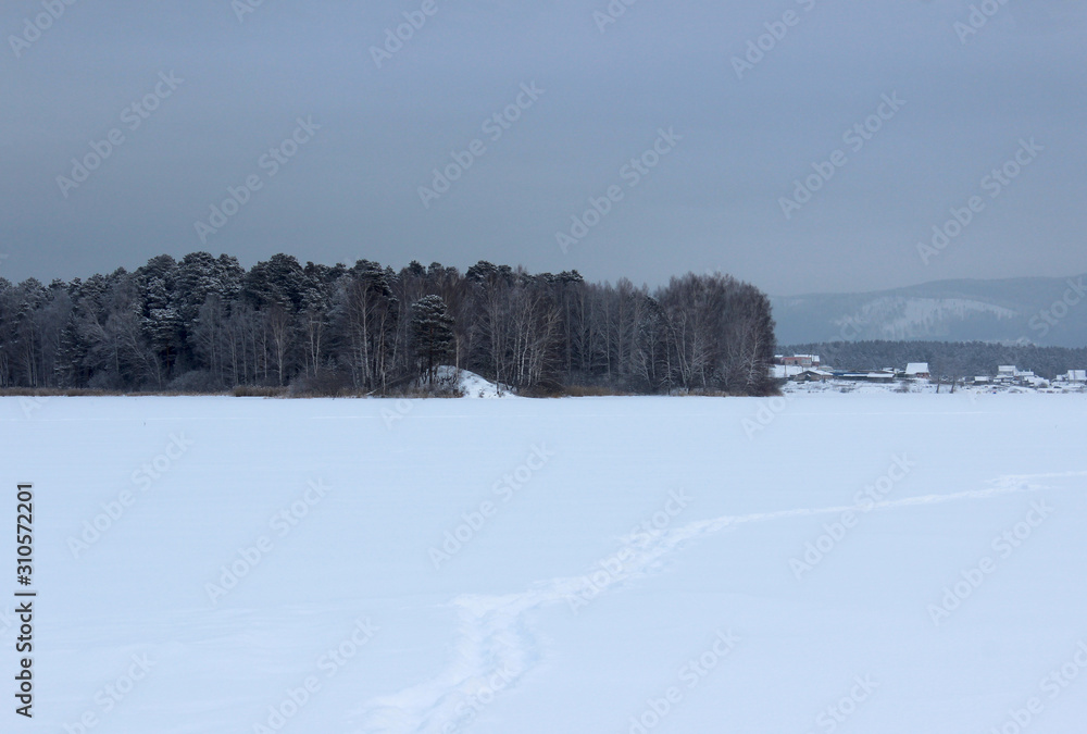 Winter landscape on the lake, forest and town houses in the background. A snow trail extending into the distance to the city. White silence, snow and sky. Winter without the sun.