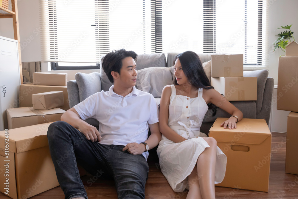 young asian couple sitting and smiling together while taking break from moving stuff in to new home with happiness. starting new life concept
