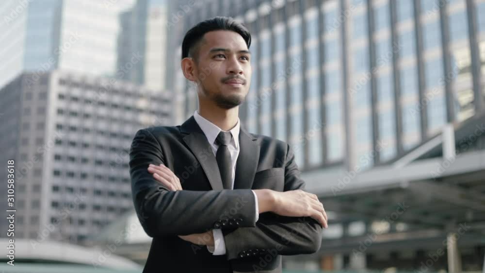 Portrait of a smiling handsome businessman in black suit thinking about future business projects while standing outside on the street near big office building urban having sunlight sunset.