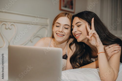 Portrait of two charming sisters having fun while talking at a laptop online laughing leanin on a bed in pijama at home. photo