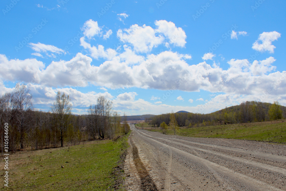 Beautiful spring day after rain in the countryside. Blue sky in the clouds and a road stretching into the distance. Early spring. The first leaves on the trees, green grass erupts. Selective focus