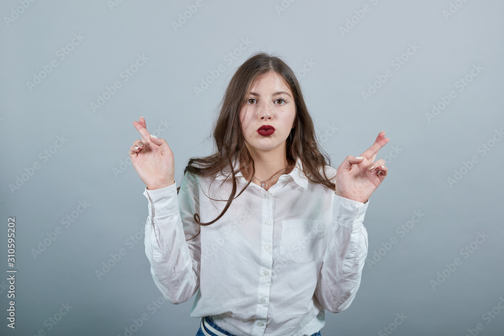 Caucasian young brunette woman over isolated gray background wearing casual white shirt, keeping fingers crossed, make wish, winks. People lifestyle concepte. Caucasian young brunette woman over isolated gray background wearing casual white shirt, keeping fingers crossed, make wish, winks. People lifestyle concepte.