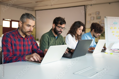Line of focused coworkers sitting at meeting table with laptops. Business colleagues in casual working together in contemporary office space. Teamwork concept