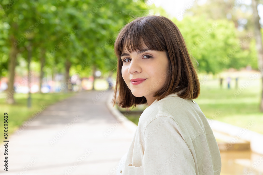 Attractive young woman smiling at camera outdoors. Portrait of beautiful cheerful young woman standing and looking at camera in park. Facial expression concept Attractive young woman smiling at camera outdoors. Portrait of beautiful cheerful young woman standing and looking at camera in park. Facial expression concept