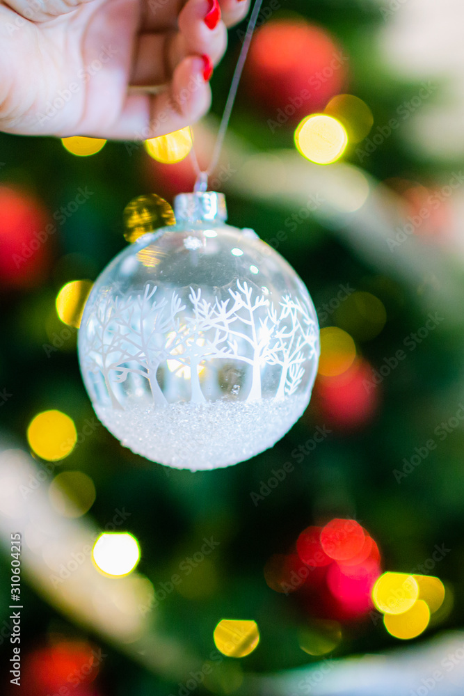 Fototapeta premium Christmas, decoration, holidays and people concept - close up of woman hand holding christmas ball over living room and tree background