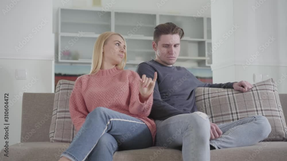 Young Caucasian woman pushing man's shoulder and giving him little finger to make peace. Young argued family sitting on sofa at home. Happiness, problems, communication, fun. Young Caucasian woman pushing man's shoulder and giving him little finger to make peace. Young argued family sitting on sofa at home. Happiness, problems, communication, fun.