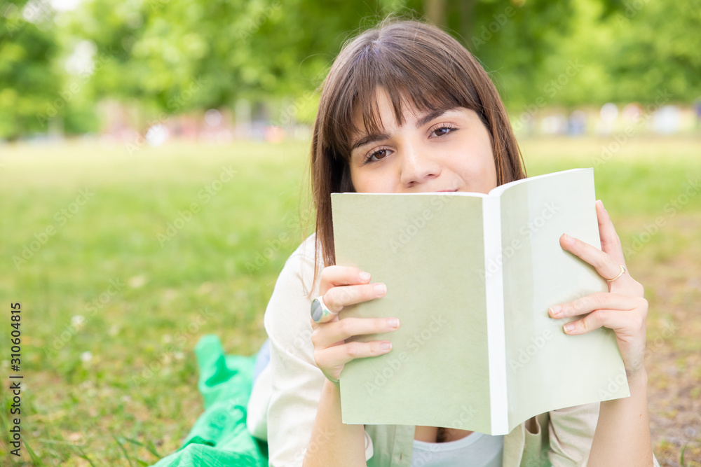 Young woman reading book in park. Attractive young woman lying on lawn with book and looking at camera in park. Education concept Young woman reading book in park. Attractive young woman lying on lawn with book and looking at camera in park. Education concept