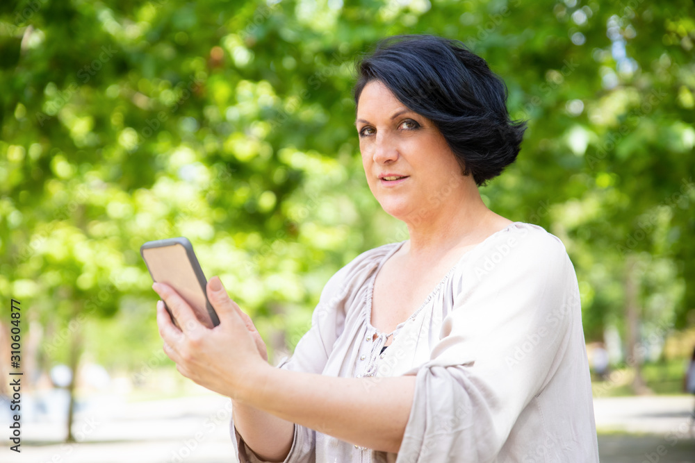 Woman holding smartphone and looking at camera. Focused middle aged woman using mobile phone in park. Technology concept