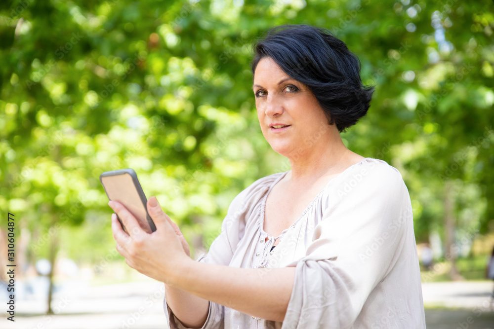 Woman holding smartphone and looking at camera. Focused middle aged woman using mobile phone in park. Technology concept Woman holding smartphone and looking at camera. Focused middle aged woman using mobile phone in park. Technology concept