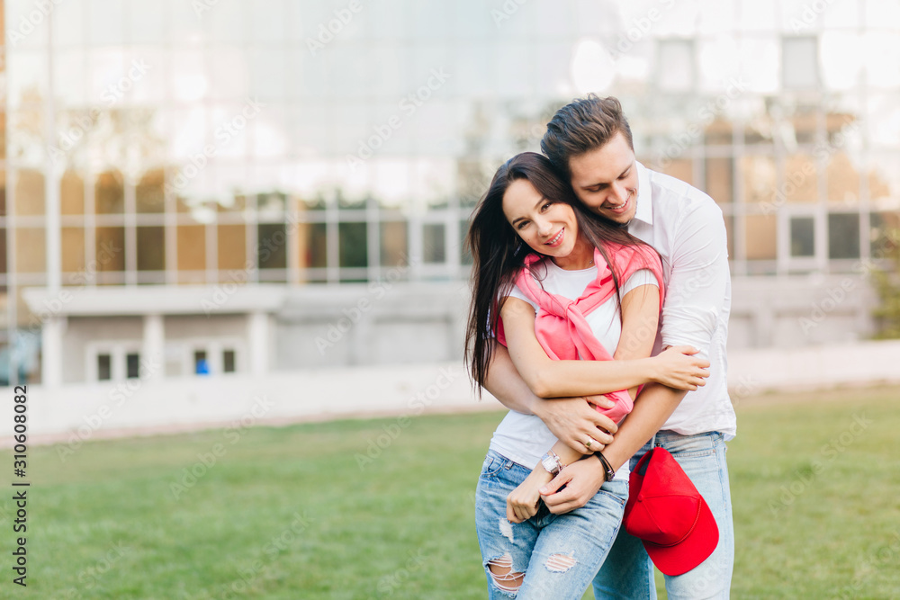 Shapely girl in blue jeans in wristwatch huggs with husband near lawn in front of building. Happy young man with red cap on pants posing with attractive woman.
