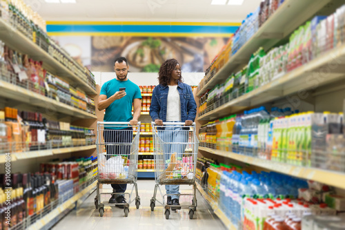Concentrated young couple walking in aisle with alcohol drinks. African American people making purchases according to shopping list. Shopping concept
