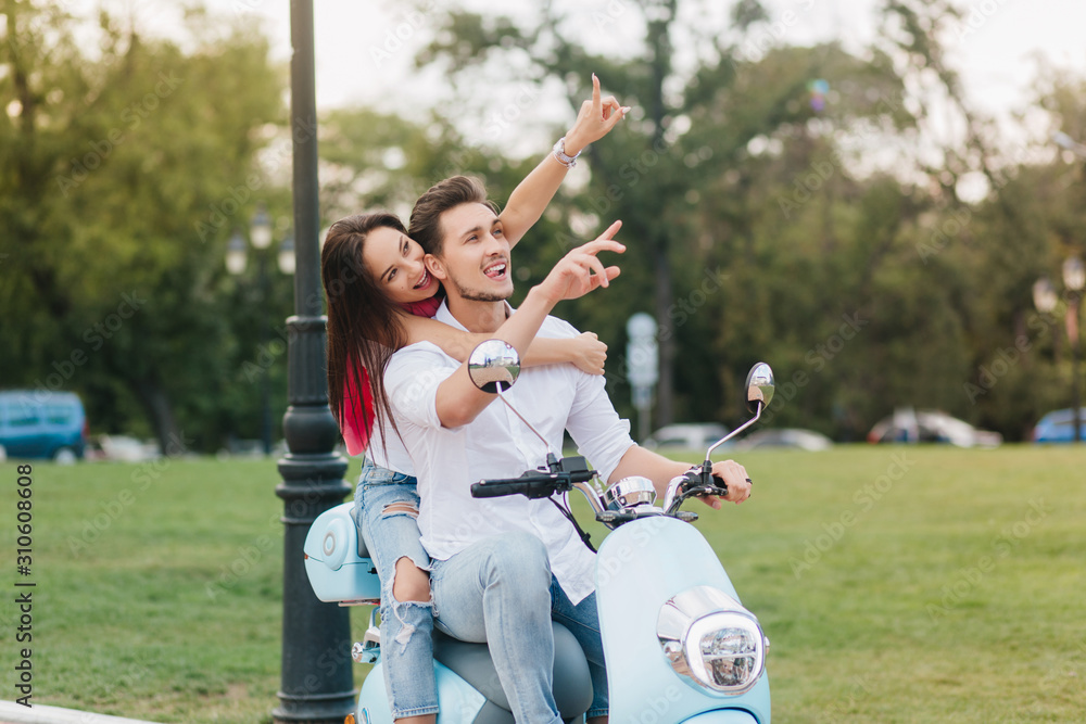 Pretty long-haired young woman enjoying trip on scooter with boyfriend. Blissful loving couple riding around park and smiling in good summer weekend.