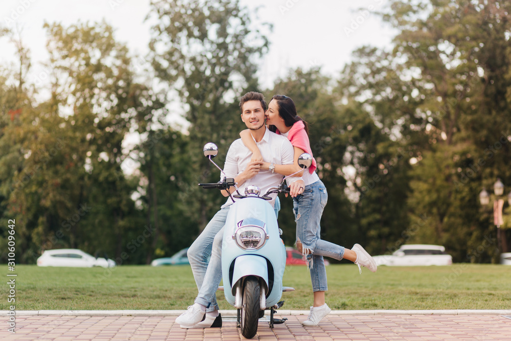 Glad girl in trendy jeans kissing boyfriend on scooter on blur nature  background. Caucasian man in white clothes enjoying outdoor date with  romantic young lady, standing beside him. Stock Photo | Adobe