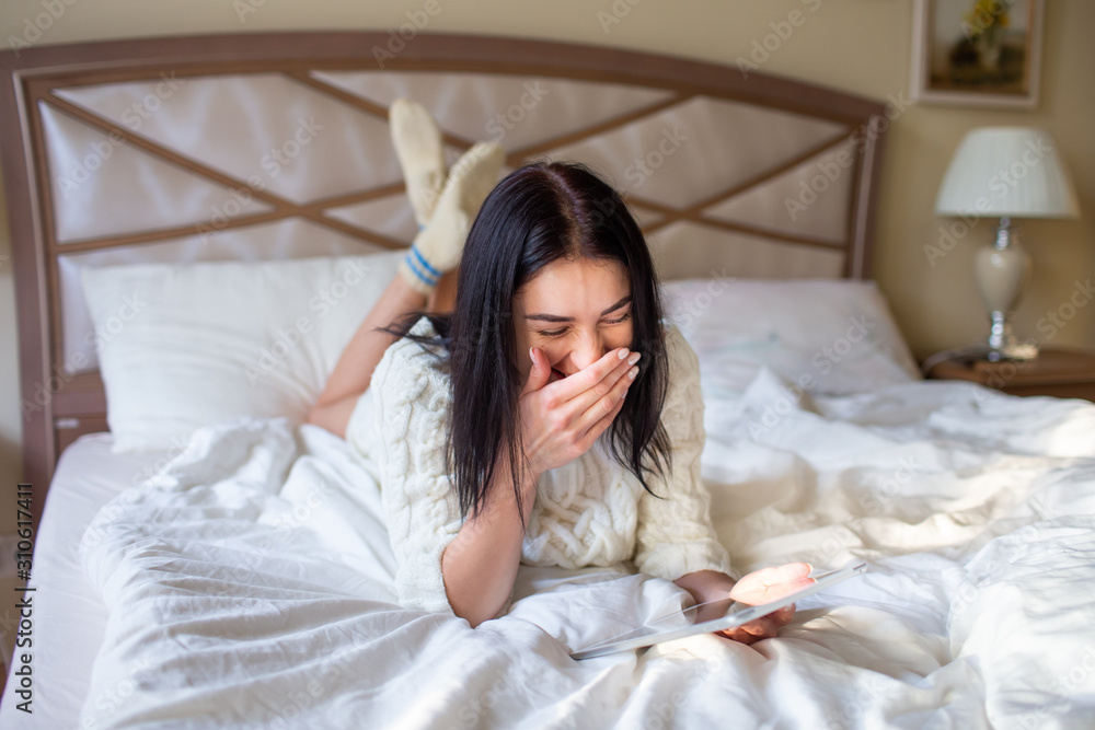 Pretty brunette girl in white knitted dress lying on bed with a tab in and hands and laughing covering her mouth with a hand Pretty brunette girl in white knitted dress lying on bed with a tab in and hands and laughing covering her mouth with a hand