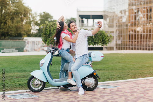 Smiling couple in jeans sitting on scooter with lawn on background. Cheerful european man making selfie on moped with his black-haired girlfriend in good day.