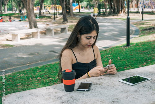 One long hair Indian female holding mobile phone in outdoor park. On table is a touch screen tablet and an eco-friendly reusable cup.
