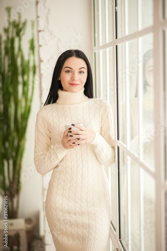Portrait of a brunette girl at the window with reflection closeup. Young woman in a white knitted dress on a background of a large window and copy space.