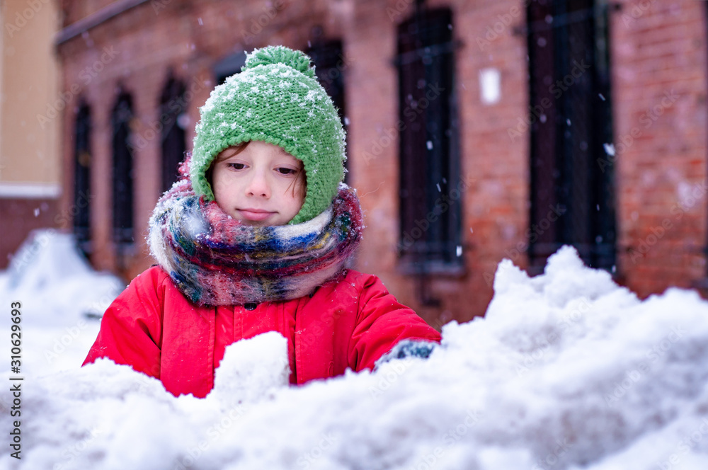 Pensive cute child playing snowballs outdoors in a snowy city in solitude. Winter leisure and children's games. Healthy lifestyle and children's activities. Girl boy in winter clothes. Pensive cute child playing snowballs outdoors in a snowy city in solitude. Winter leisure and children's games. Healthy lifestyle and children's activities. Girl boy in winter clothes.