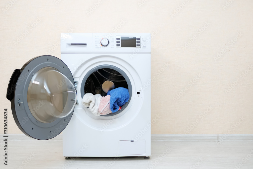 modern washing machine with heap of laundry in drum and open door stands on wooden floor near beige wall