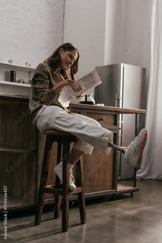 Smiling young woman with leg prosthesis reading newspaper at kitchen