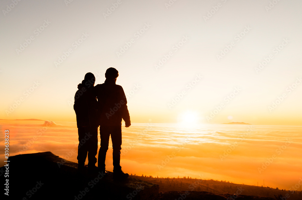 Silhouette of two friends man standing on a rock at sunrise on the fog of success and serenity.