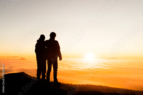 Silhouette of two friends man standing on a rock at sunrise on the fog of success and serenity.